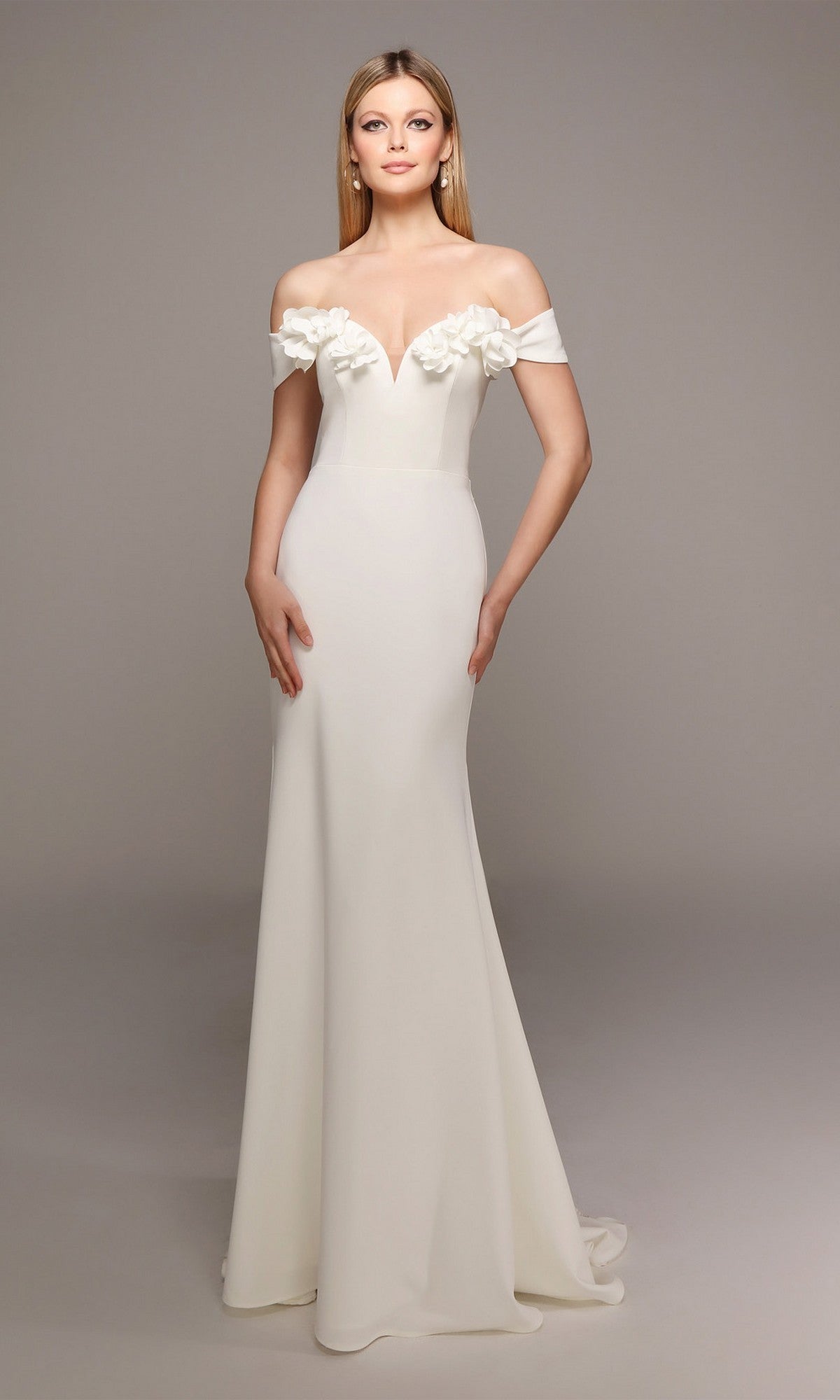 A woman wearing the Alyce 7062 long formal dress in ivory, featuring an off-the-shoulder neckline and 3D floral bodice accents, stands against a plain gray background.